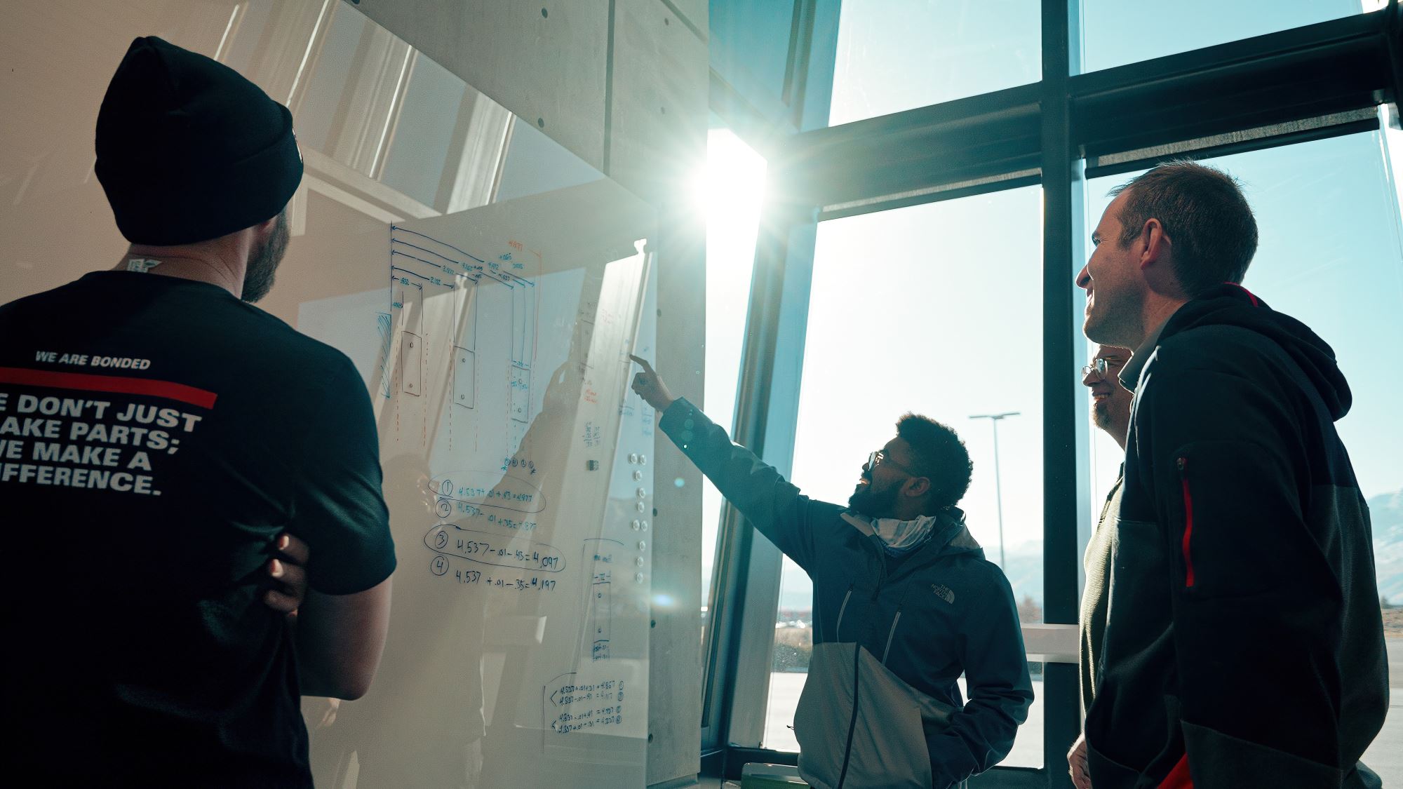 Click Bond engineers collaborating on adhesive bonding solutions, reviewing technical drawings and design data on a whiteboard inside a sunlit engineering facility.
